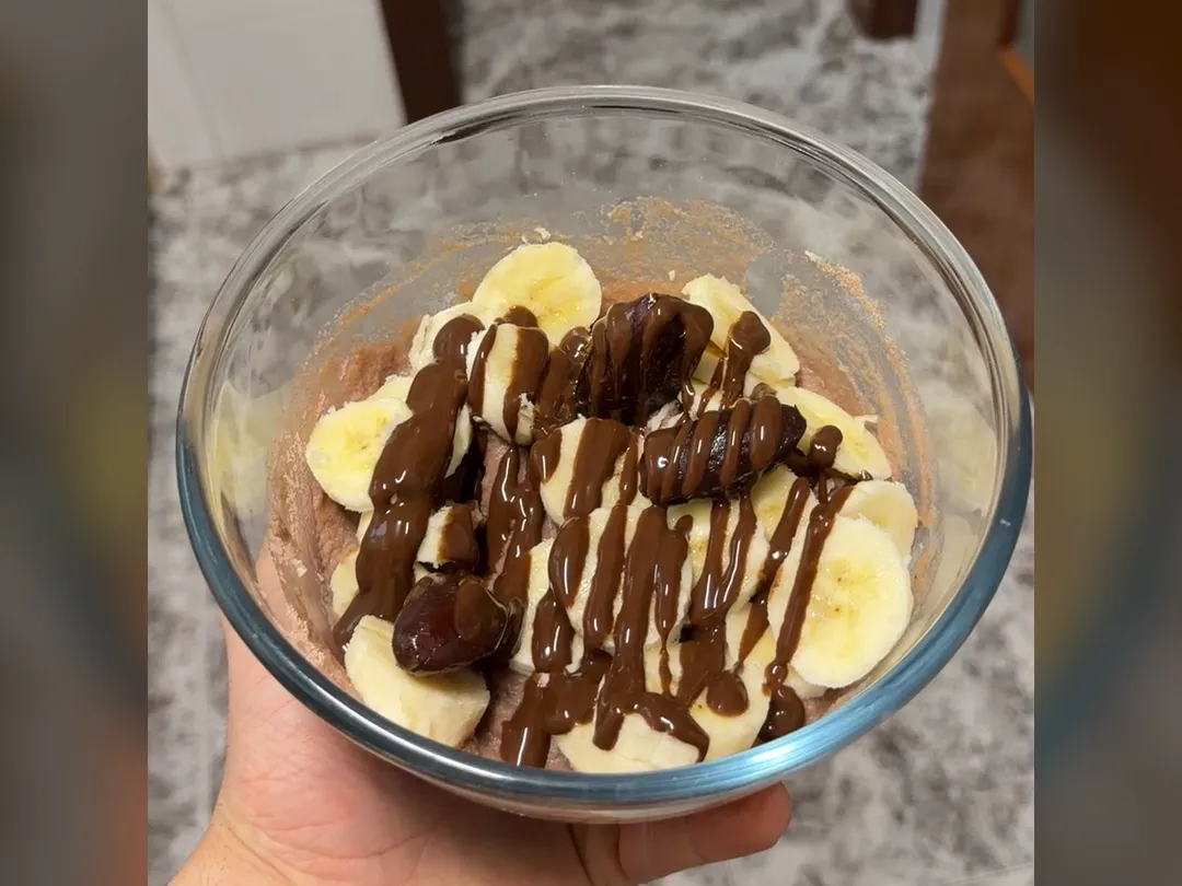 Bol de crema de arroz con plátano en rodajas, dátiles y sirope de chocolate, en la mano