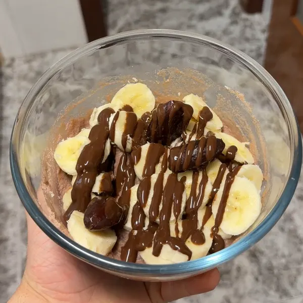Bol de crema de arroz con plátano en rodajas, dátiles y sirope de chocolate, en la mano
